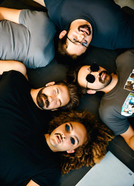three guys and one girl lay face down. they are wearing black T-shirts. One guy with a beard, the second guy with a Mustache, the third with a fluffy haircut, a girl with a bob